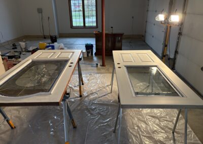 Two doors with glass panels rest on sawhorses for painting or repairs in a garage. Plastic sheets cover the floor, tools and supplies are scattered, and a work light illuminates the scene—a typical setup for a residential painting Upstate New York project.