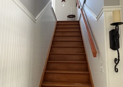 A wooden staircase with white wainscoting on the walls leads up to a landing. On the landing, there is a small table with a decorative white branch and a wall-mounted star ornament above it.